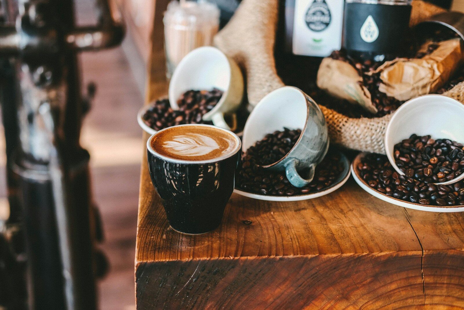 Latte art adorns a black cup, resting on a wooden table surrounded by scattered coffee beans and overturned cups, highlighting a cozy café ambiance.