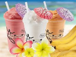Colorful tropical smoothies in decorative cups, adorned with small umbrellas, surrounded by bananas and flowers on a beach backdrop.