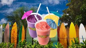 Three colorful smoothies topped with umbrellas, set against a vibrant backdrop of palm trees and a colorful surfboard fence, evoking a tropical vibe.