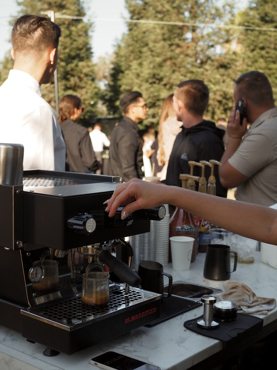 A hand adjusts a coffee machine in a bustling outdoor setting, with guests mingling in the background, highlighting a lively event atmosphere.