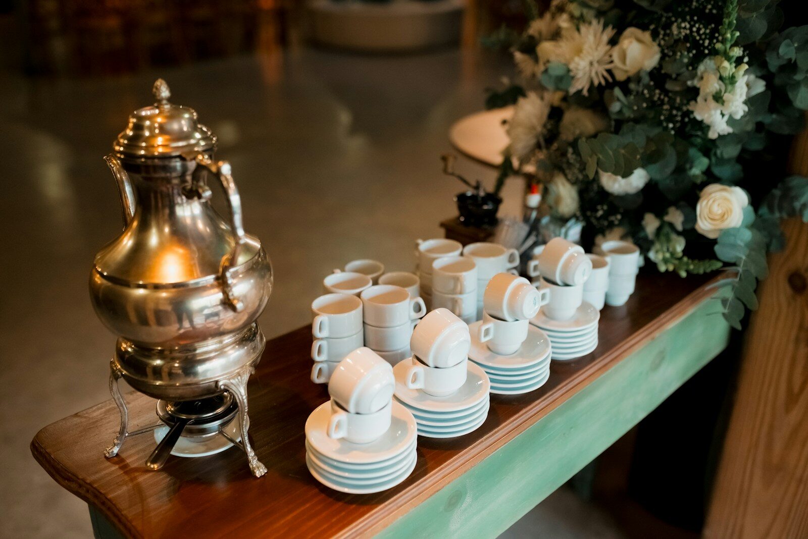 A silver samovar with white coffee cups stacked on saucers, set on a wooden table adorned with flowers, ready for a gathering.