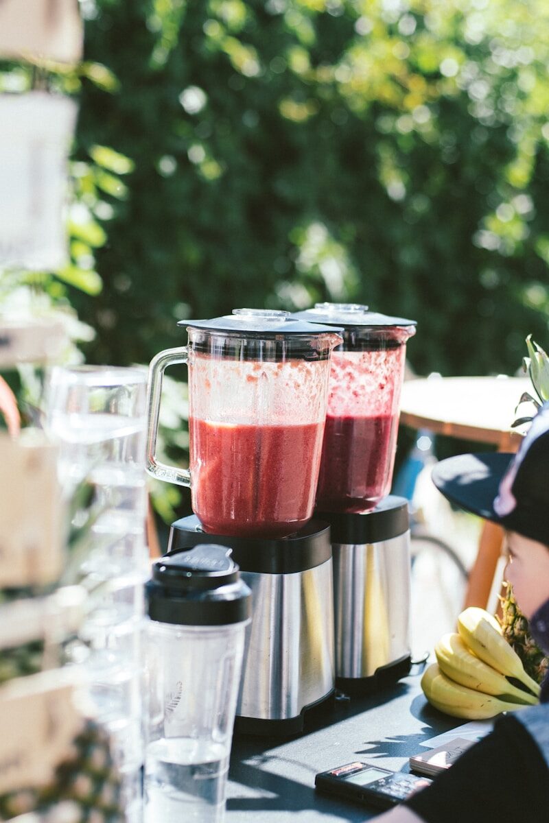Two blenders filled with vibrant red smoothies sit on a table, surrounded by glass cups and bananas, indicating a refreshing drink stand setup.